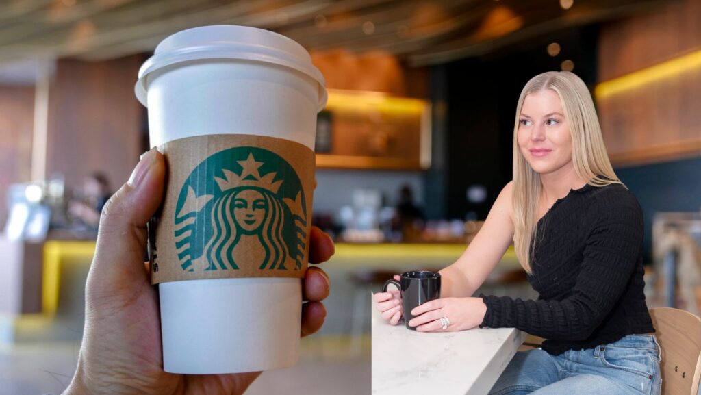 Close-up of a white Starbucks takeaway cup with sleeve in the foreground, being offered toward a smiling woman at a cafe counter in the background.