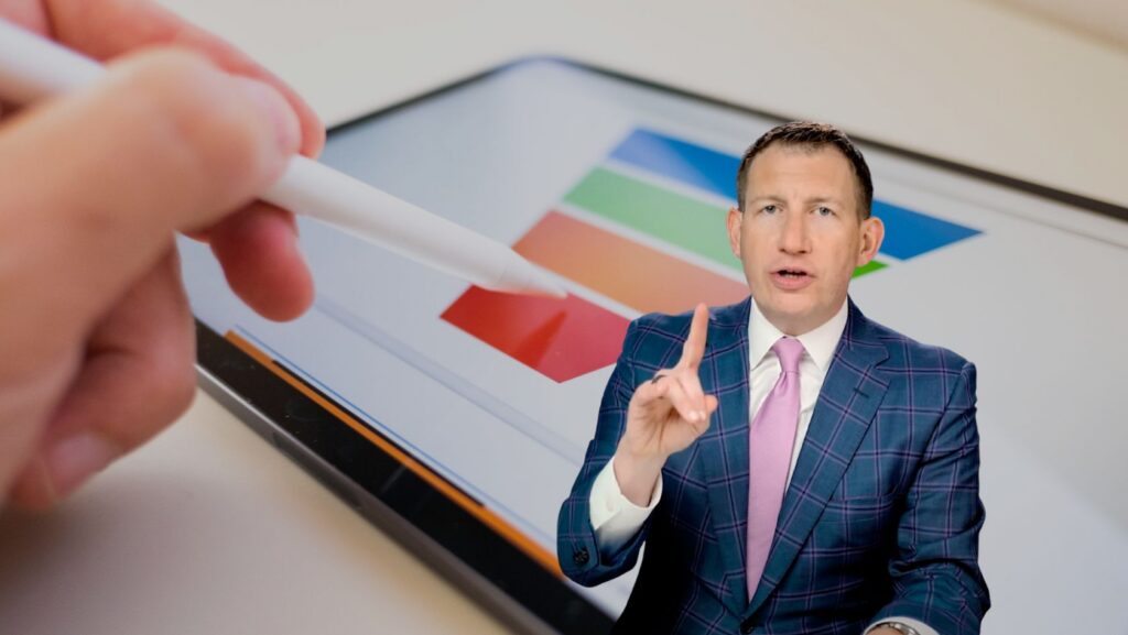 Man in a blue plaid suit and pink tie gesturing while presenting, with a tablet showing color swatches in the background.