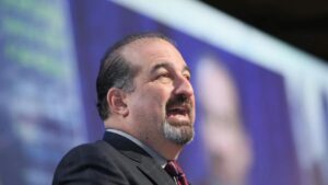 Man in a suit giving a speech on a stage with a purple background.