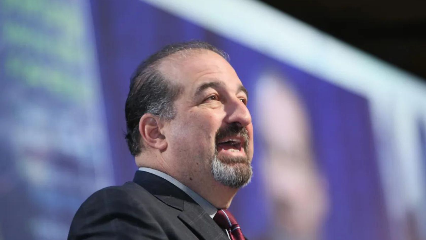 Man in a suit giving a speech on a stage with a purple background.