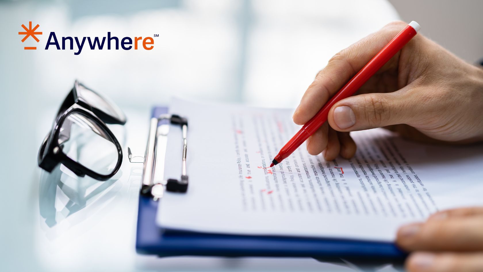 Hand signing a contract with a red pen on a clipboard; glasses sit nearby with the Anywhere logo in the top left corner.