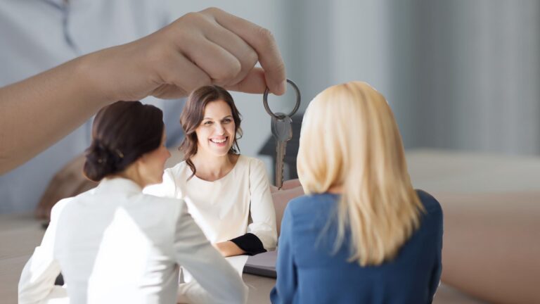 Smiling woman at a table with two colleagues as a giant hand hands her a key, symbolizing receiving the keys to a new home or office.