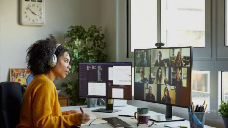 Woman wearing headphones works at a desk with two monitors, one showing a video conference grid and the other sparse documents/code.