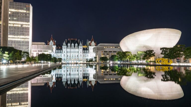 Night cityscape: a glass office tower, historic white buildings, and a large curved white museum, all reflected in a calm pool of water.
