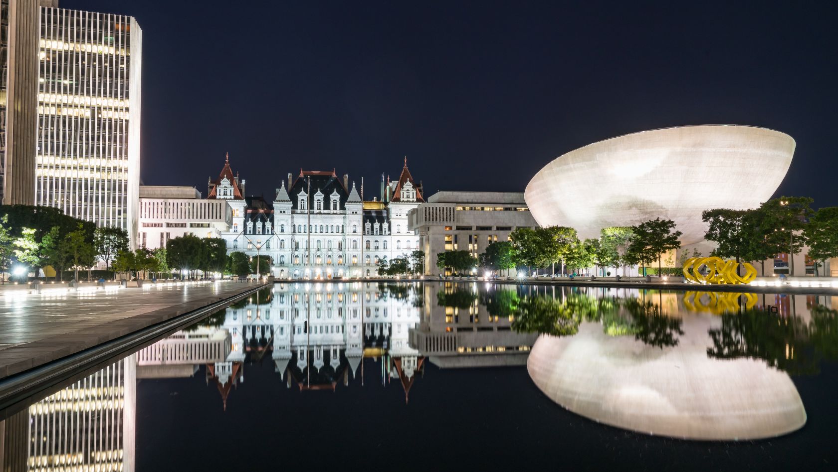 Night cityscape: a glass office tower, historic white buildings, and a large curved white museum, all reflected in a calm pool of water.