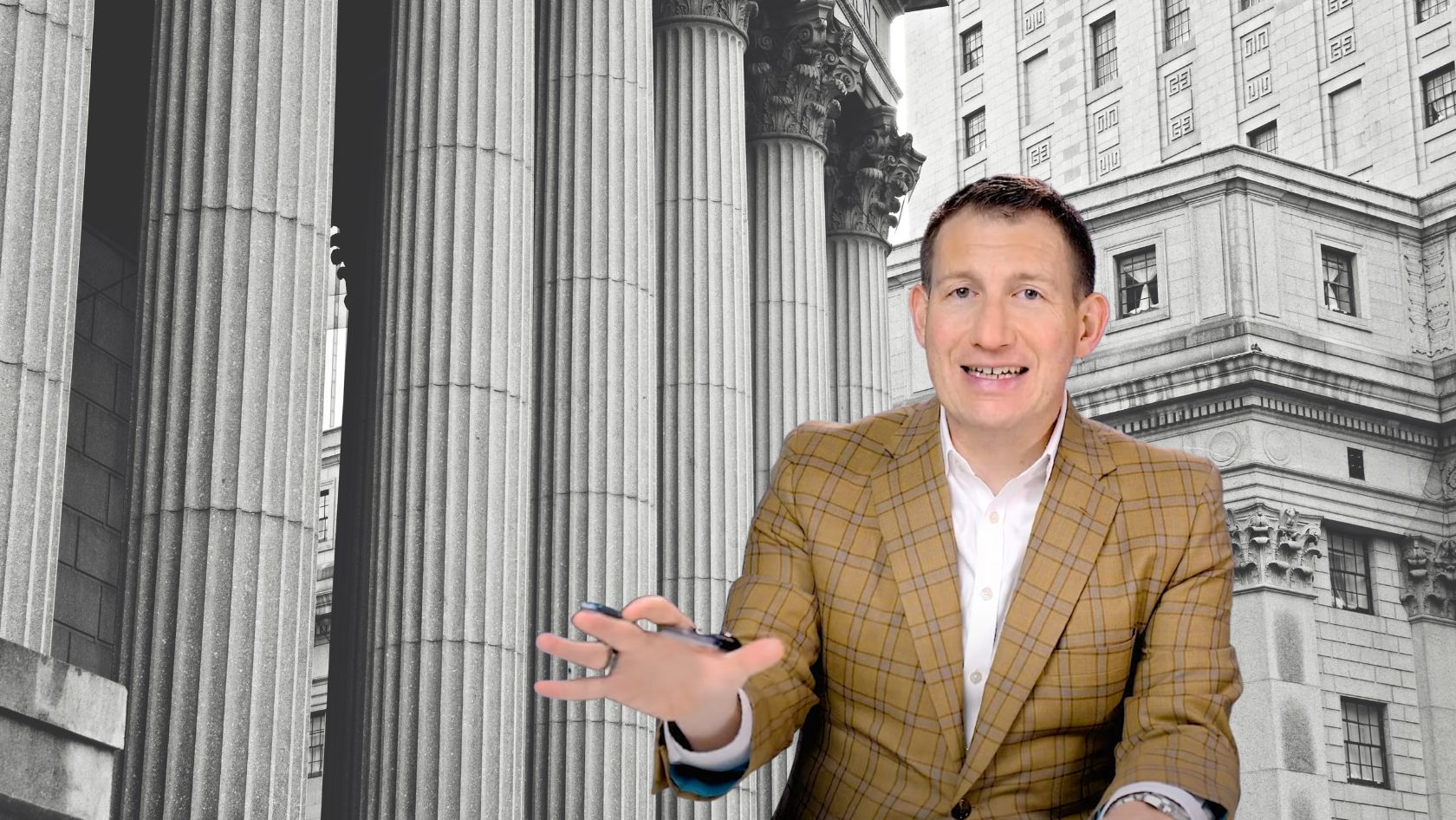 Smiling man in a tan plaid blazer reaches forward with his hand, against a backdrop of tall stone columns and a classical building behind him.