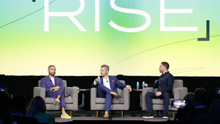 Three men in suits sit on a stage panel, each with a drink, against a large green-yellow backdrop with the word 'RISE'.