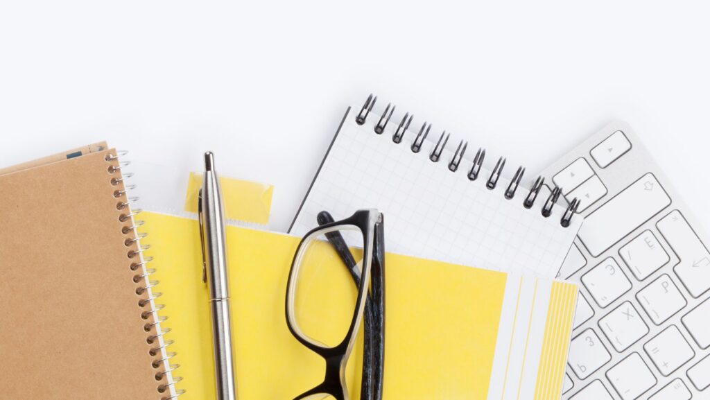 Top-down view of an office desk with spiral notebooks, yellow sticky notes, a pen, and a pair of glasses beside a white keyboard.