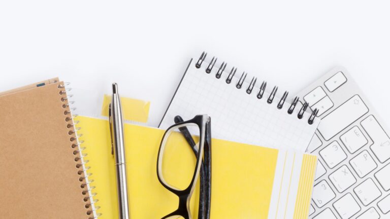 Top-down view of an office desk with spiral notebooks, yellow sticky notes, a pen, and a pair of glasses beside a white keyboard.