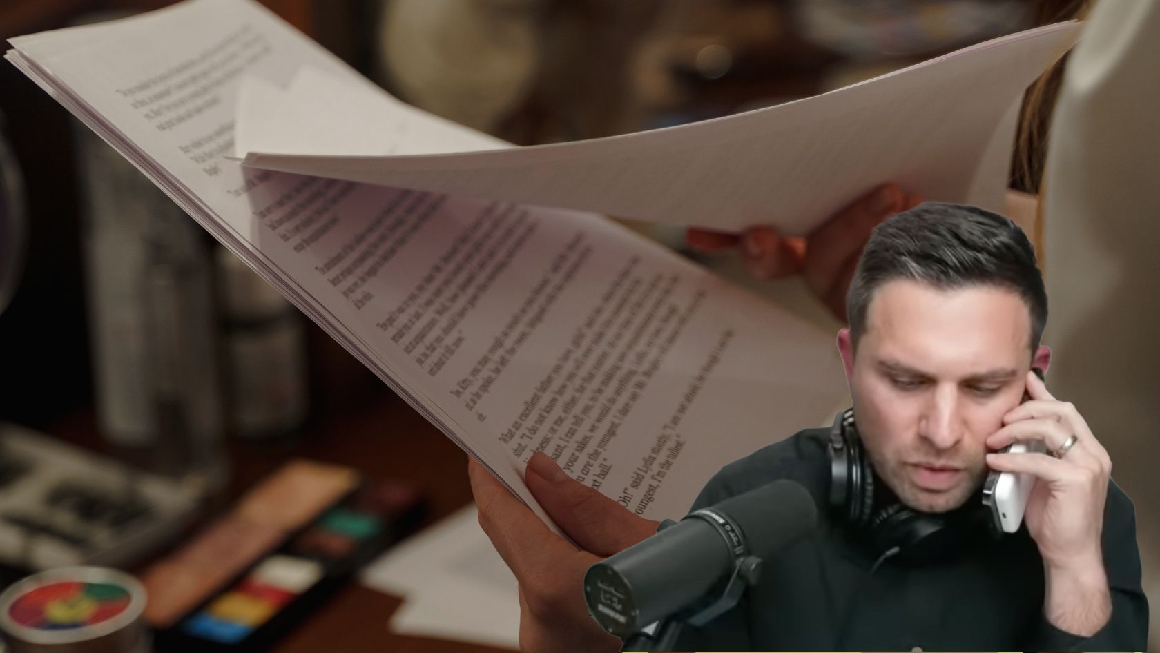 Podcaster on a microphone, talking on a phone while reviewing stacked papers in a studio setting.