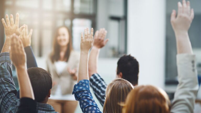 People in a meeting raising their hands to answer a question, with a speaker in the background in a bright room.