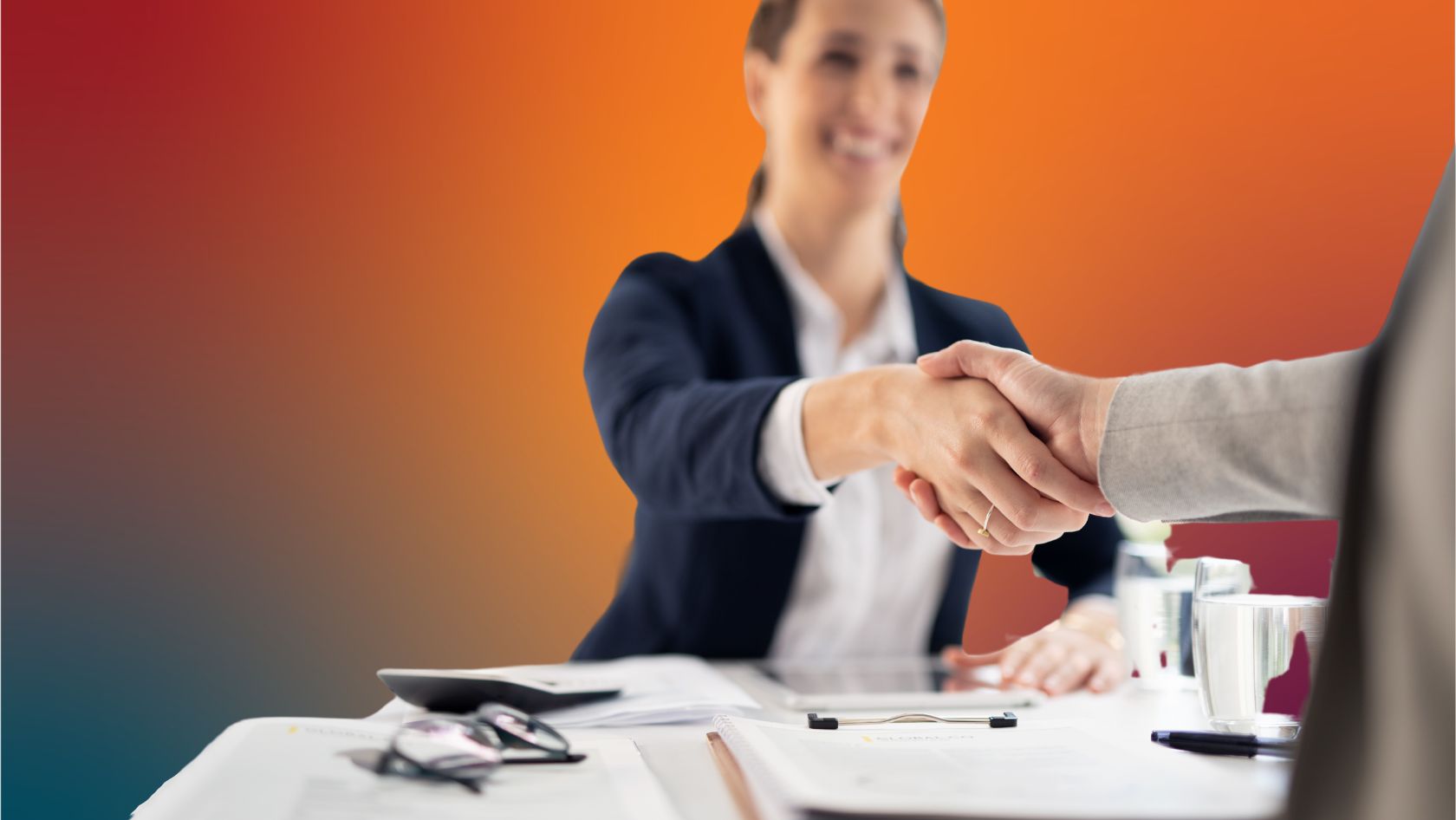Two professionals shake hands across a desk in an office, signaling an agreement or partnership.