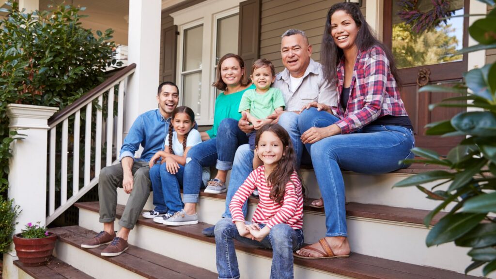 A family of six sits on the front steps of a house, smiling at the camera.