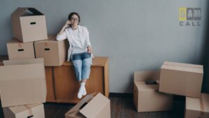 Woman sits on a wooden cabinet, talking on her phone, surrounded by stacked moving boxes in a room with a '5AM CALL' logo on the wall.