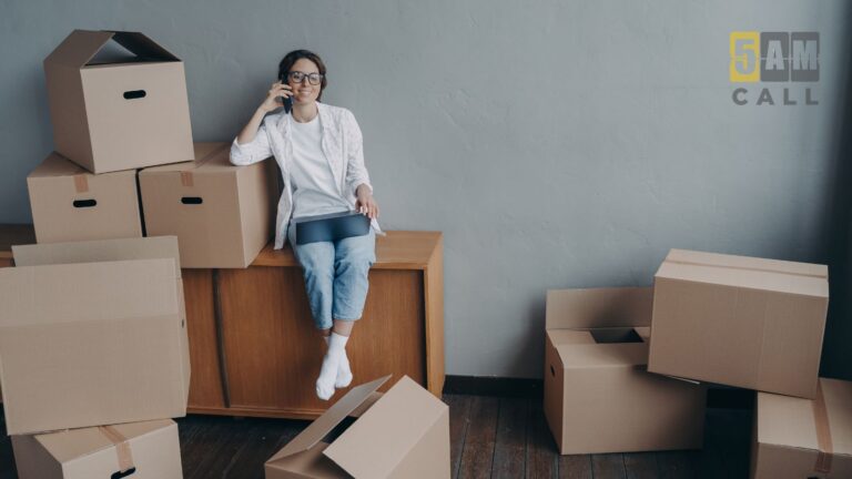 Woman sits on a wooden cabinet, talking on her phone, surrounded by stacked moving boxes in a room with a '5AM CALL' logo on the wall.