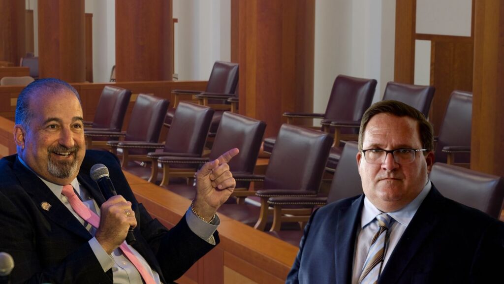Two men in suits sit in a wood-paneled hearing room; one holds a microphone and points as he speaks, the other listens nearby in glasses.