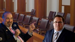 Two men in suits sit in a wood-paneled hearing room; one holds a microphone and points as he speaks, the other listens nearby in glasses.