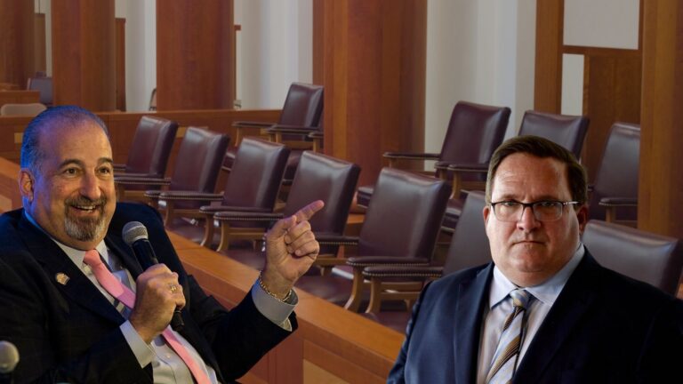 Two men in suits sit in a wood-paneled hearing room; one holds a microphone and points as he speaks, the other listens nearby in glasses.