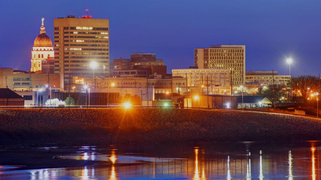 Night city skyline at dusk with illuminated buildings along a river and reflections in the water, left by a domed historic tower.
