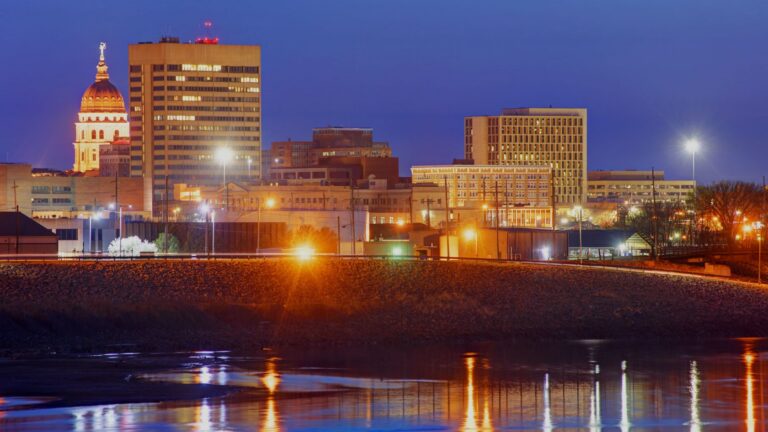 Night city skyline at dusk with illuminated buildings along a river and reflections in the water, left by a domed historic tower.