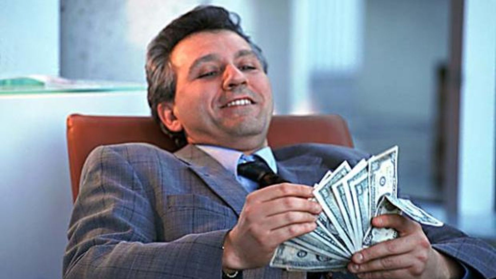 Smiling businessman in a gray suit sits in an office, counting a stack of dollar bills.