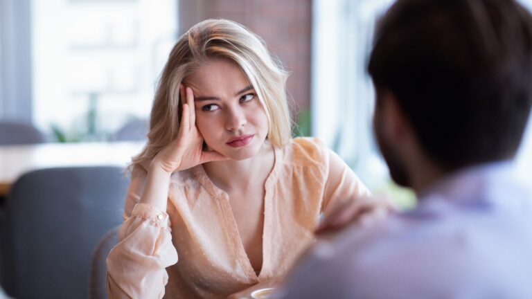 Blonde woman resting her head on her hand, listening thoughtfully to a man opposite her in a bright setting.
