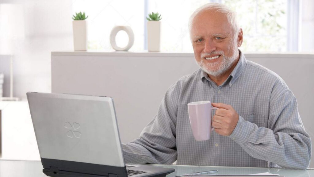 Smiling older man with white hair and beard sits at a desk with a laptop, holding a purple mug.