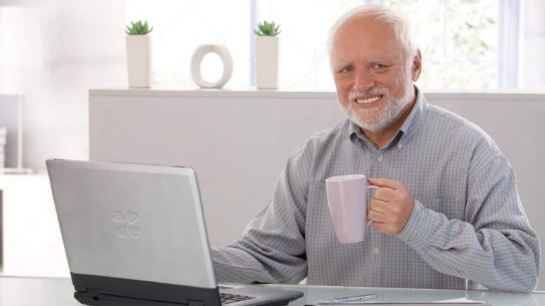Smiling older man with white hair and beard sits at a desk with a laptop, holding a purple mug.