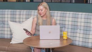 Woman with blonde hair sits in a cafe booth, using a silver laptop and checking her phone, with a yellow can on the table.