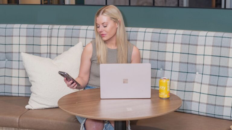 Woman with blonde hair sits in a cafe booth, using a silver laptop and checking her phone, with a yellow can on the table.