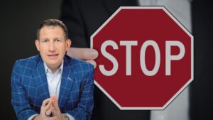 Man in a blue checkered suit sits with hands clasped beside a large red stop sign graphic