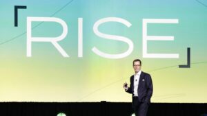 Speaker in a dark suit on a stage holding a clicker, with a large gradient backdrop displaying 'RSE'.