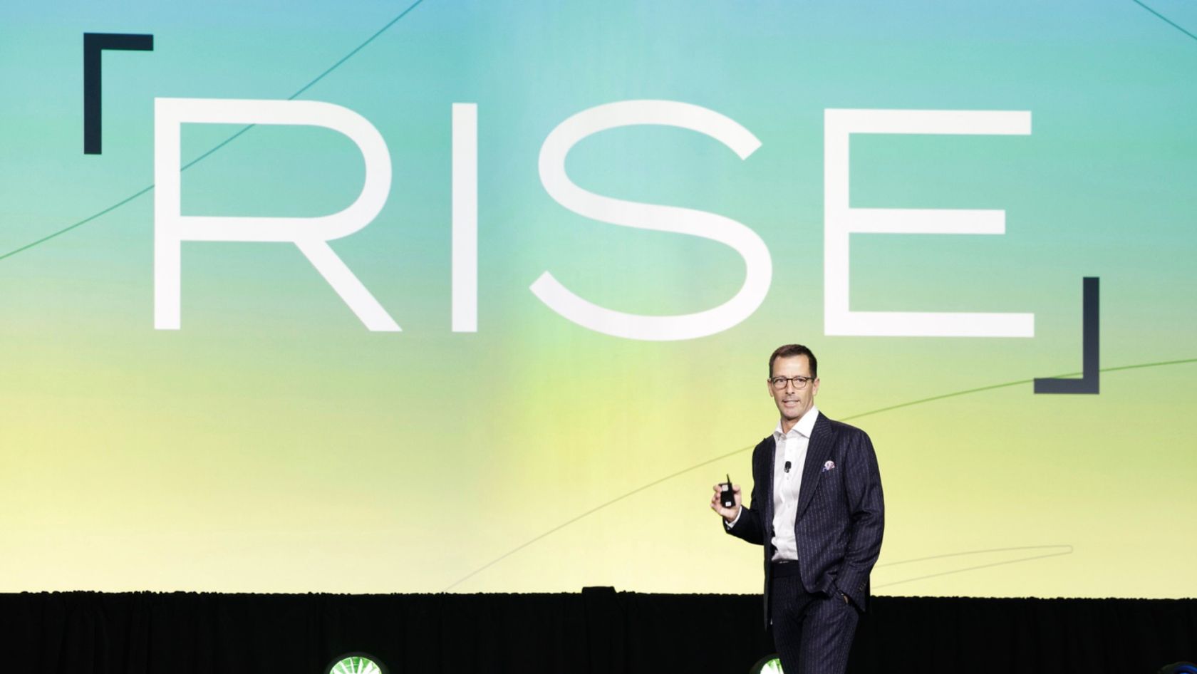 Speaker in a dark suit on a stage holding a clicker, with a large gradient backdrop displaying 'RSE'.
