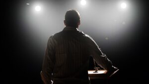 Back view of a man at a wooden lectern on a dimly lit stage with bright spotlights in the background.