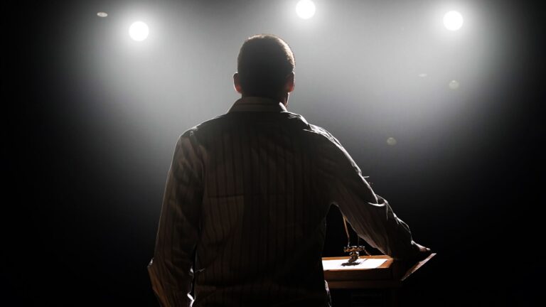 Back view of a man at a wooden lectern on a dimly lit stage with bright spotlights in the background.