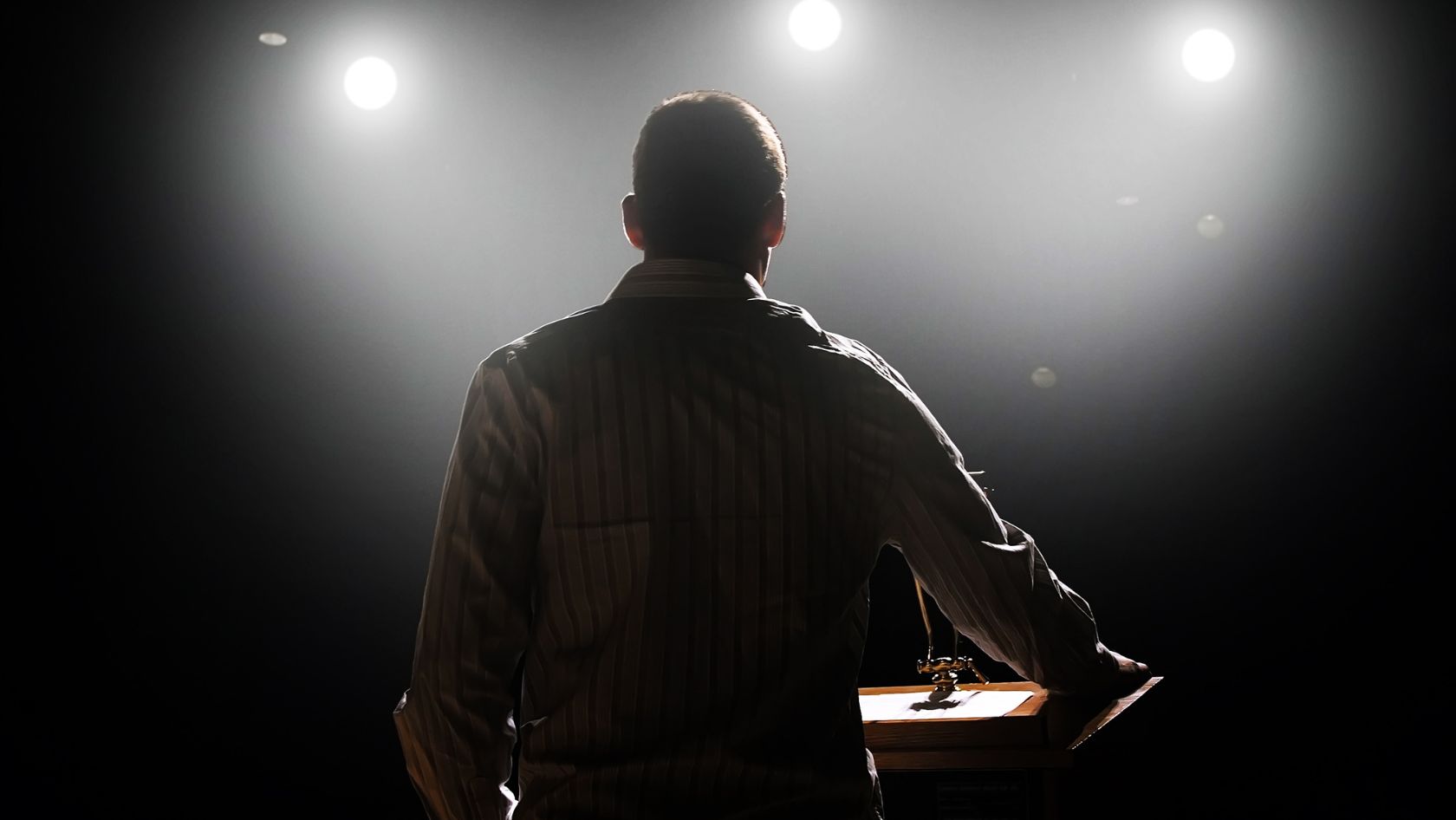 Back view of a man at a wooden lectern on a dimly lit stage with bright spotlights in the background.