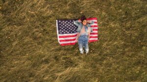 Person lying on back on an American flag spread on a grassy field, seen from above?