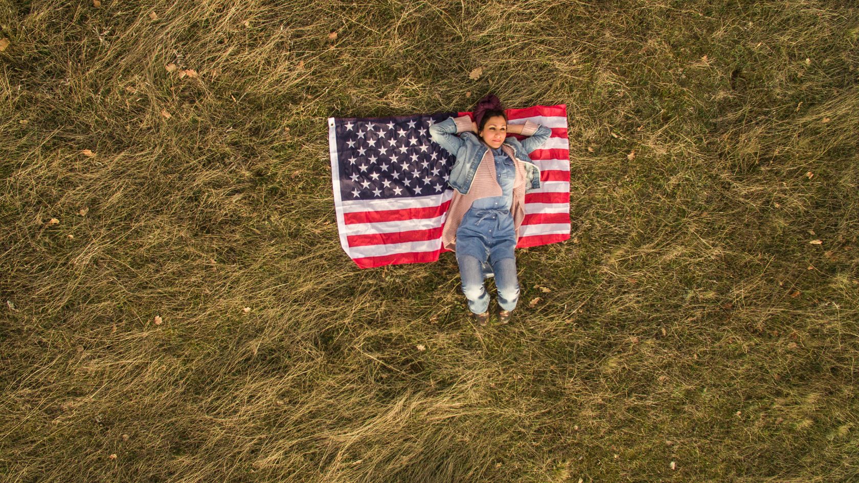 Person lying on back on an American flag spread on a grassy field, seen from above?