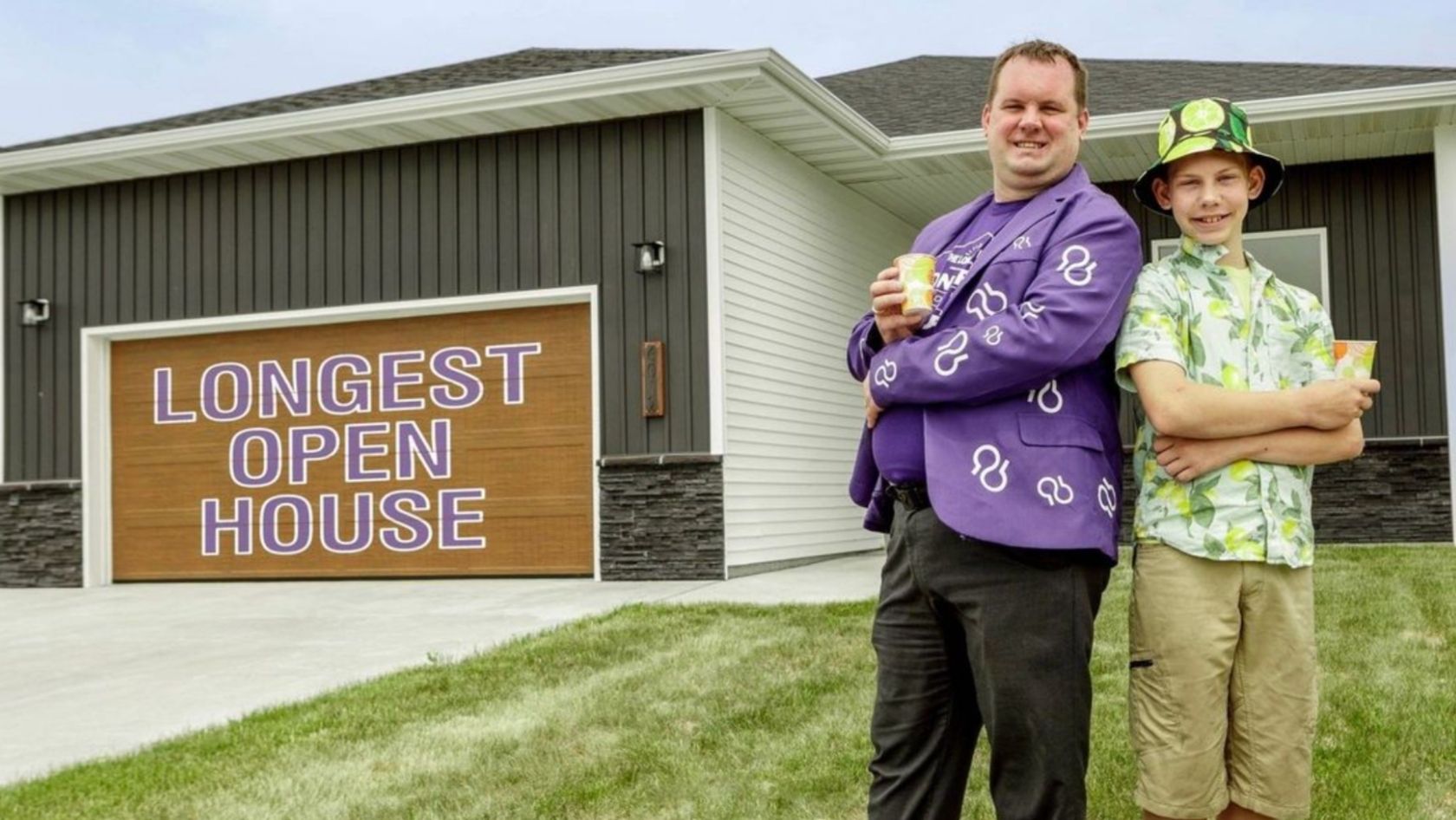 Two people pose outside a modern house next to a large sign that says LONGEST OPEN HOUSE, each holding a drink.