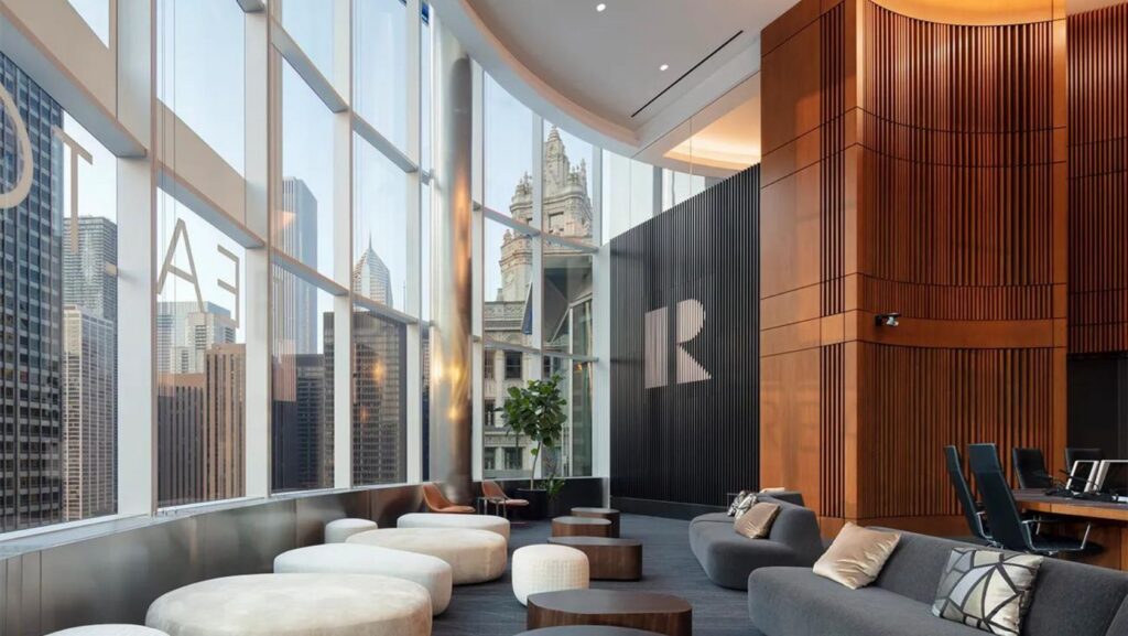 Modern corporate lounge with curved wood panel wall, gray couches, and round ottomans overlooking a glass-walled city skyline.