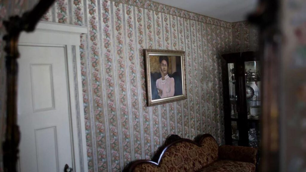 Vintage living room with floral striped wallpaper, a white paneled door on the left, and a framed portrait of a woman above a decorative patterned sofa.