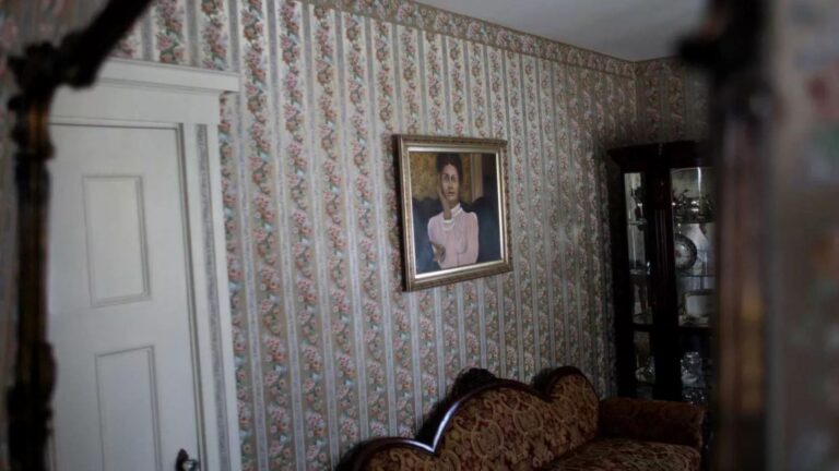 Vintage living room with floral striped wallpaper, a white paneled door on the left, and a framed portrait of a woman above a decorative patterned sofa.