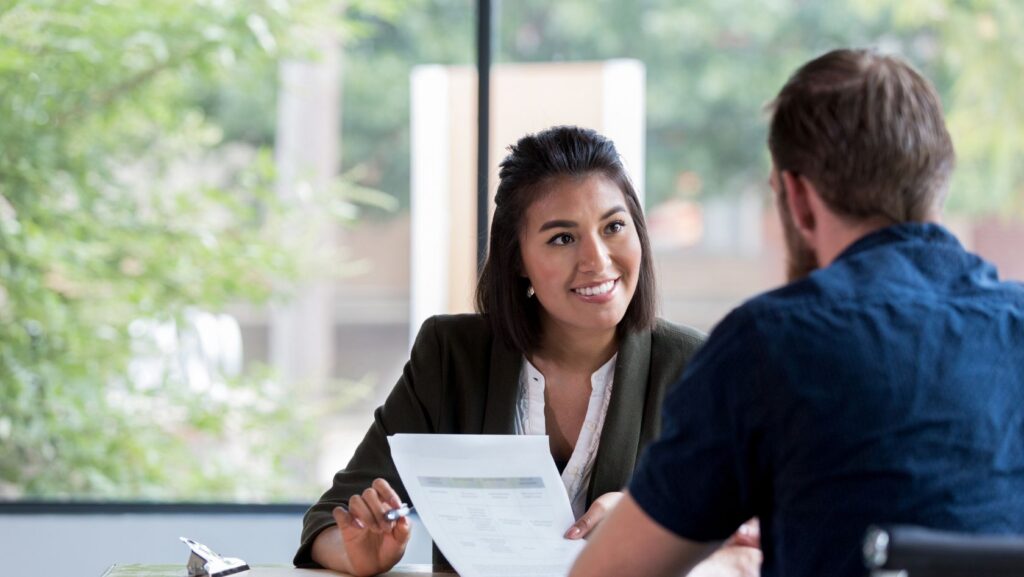 Woman in a blazer smiles while discussing a document with a man across a table in a bright office.