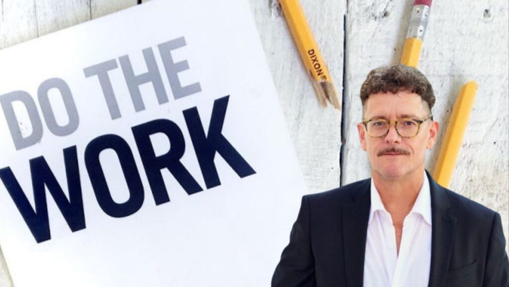 Man in a suit and glasses stands beside a large white sign that reads 'DO THE WORK' against a wooden wall with yellow pencils in the background.