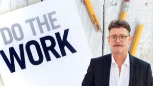 Man in a suit and glasses stands beside a large white sign that reads 'DO THE WORK' against a wooden wall with yellow pencils in the background.