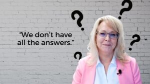 Professional woman in a pink blazer stands against a brick wall with large question marks; quote reads: We don’t have all the answers.