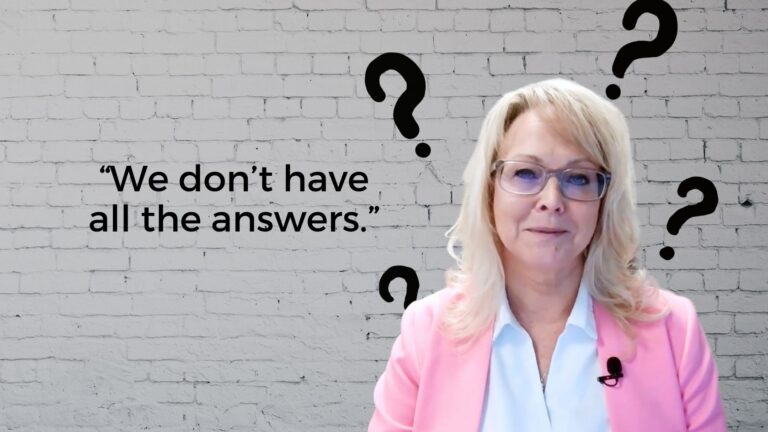Professional woman in a pink blazer stands against a brick wall with large question marks; quote reads: We don’t have all the answers.