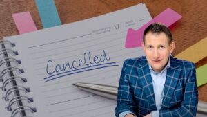 Notebook page shows 'Cancelled' written in blue with a silver pen nearby; man in a blue plaid blazer sits in the foreground.