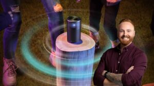 Smiling man with folded arms beside a portable speaker on a tree stump, surrounded by colorful light rings and silhouettes of people in the background.