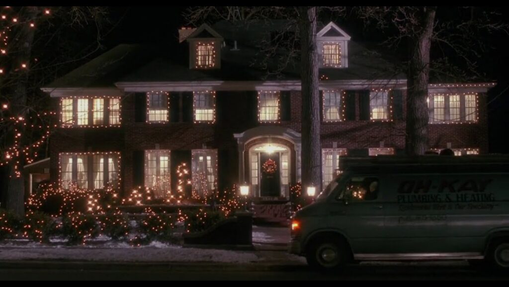 Two-story brick house at night adorned with warm white Christmas lights on windows, trees, and shrubs; a service van is parked in front.